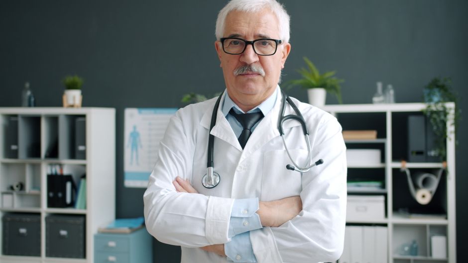 Doctor standing confidently in medical office with stethoscope, arms crossed.
