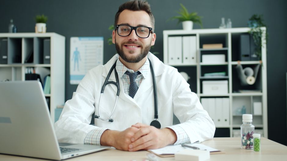Friendly male doctor with stethoscope sitting at office desk in clinic.